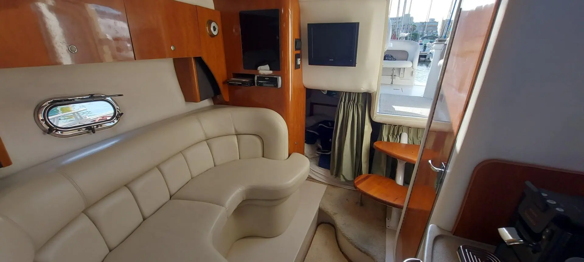 Four Winns V 288 — Salon intérieur avec banquette en U, hublot et accès vers le cockpit.