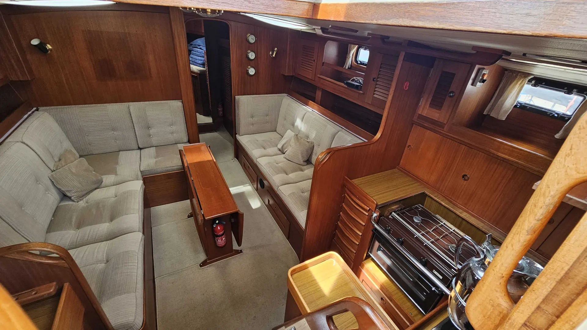 Hallberg Rassy 42 — Vue du salon intérieur en bois avec banquettes, table et cuisinière.