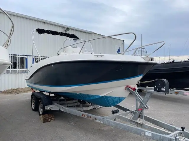Quicksilver 555 Open — Bateau à moteur blanc et bleu sur remorque, photographié de trois quarts à terre.