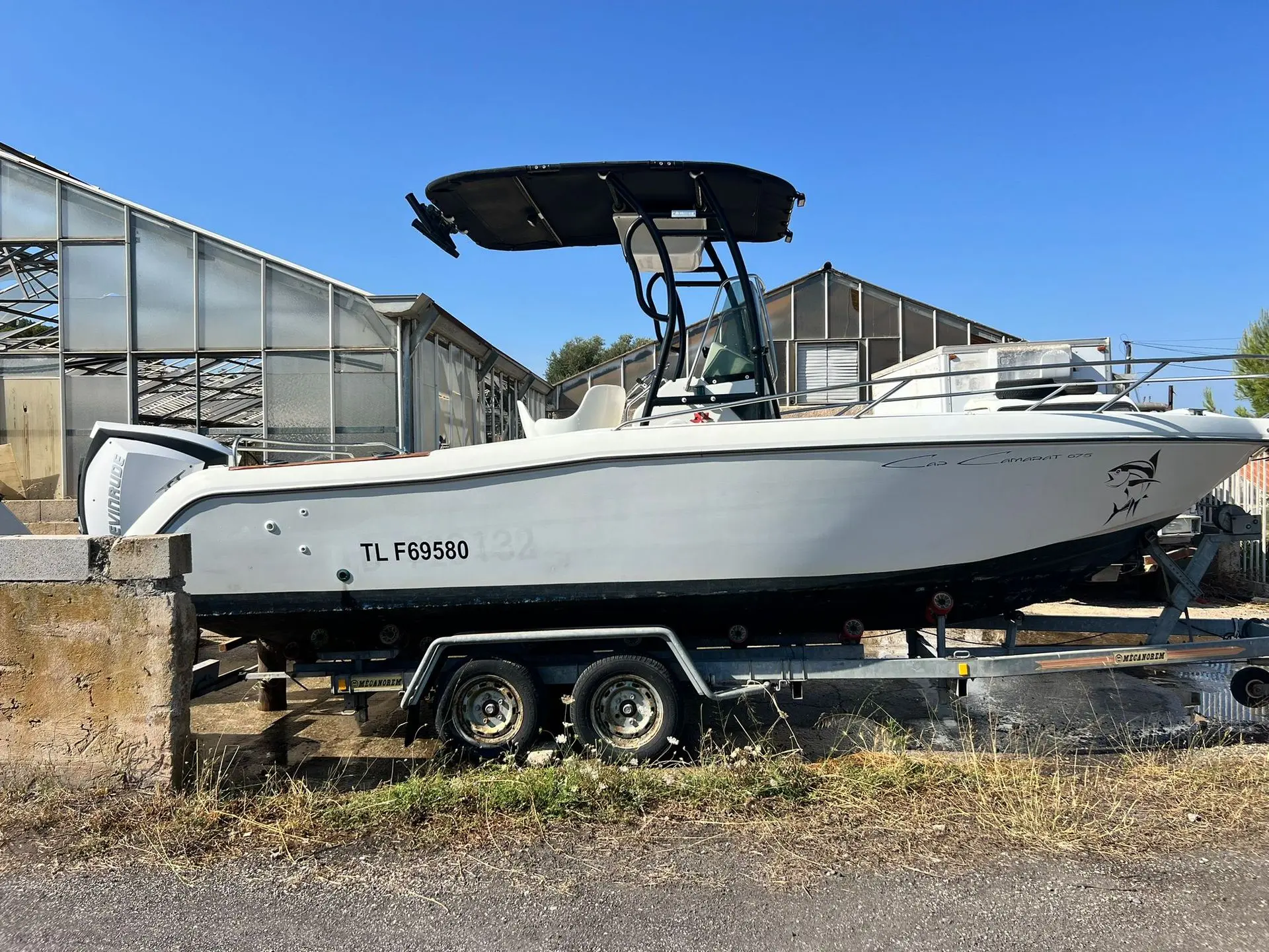 Jeanneau Cap Camarat 675 — Bateau à moteur blanc vu de profil, posé sur une remorque devant des serres.