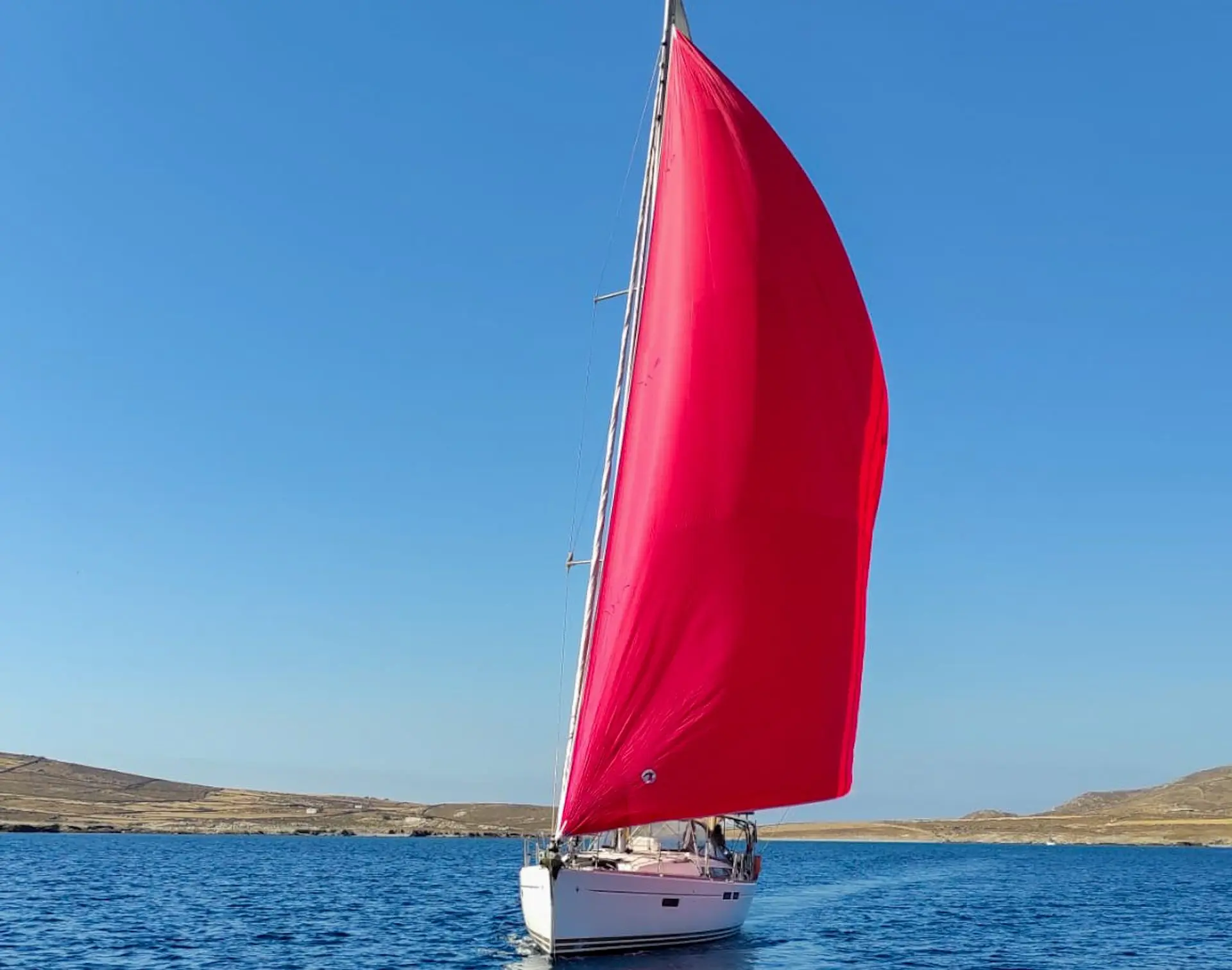 Jeanneau Sun Odyssey 469 — Voilier avançant sur une mer calme, spi rouge déployé devant un paysage côtier.