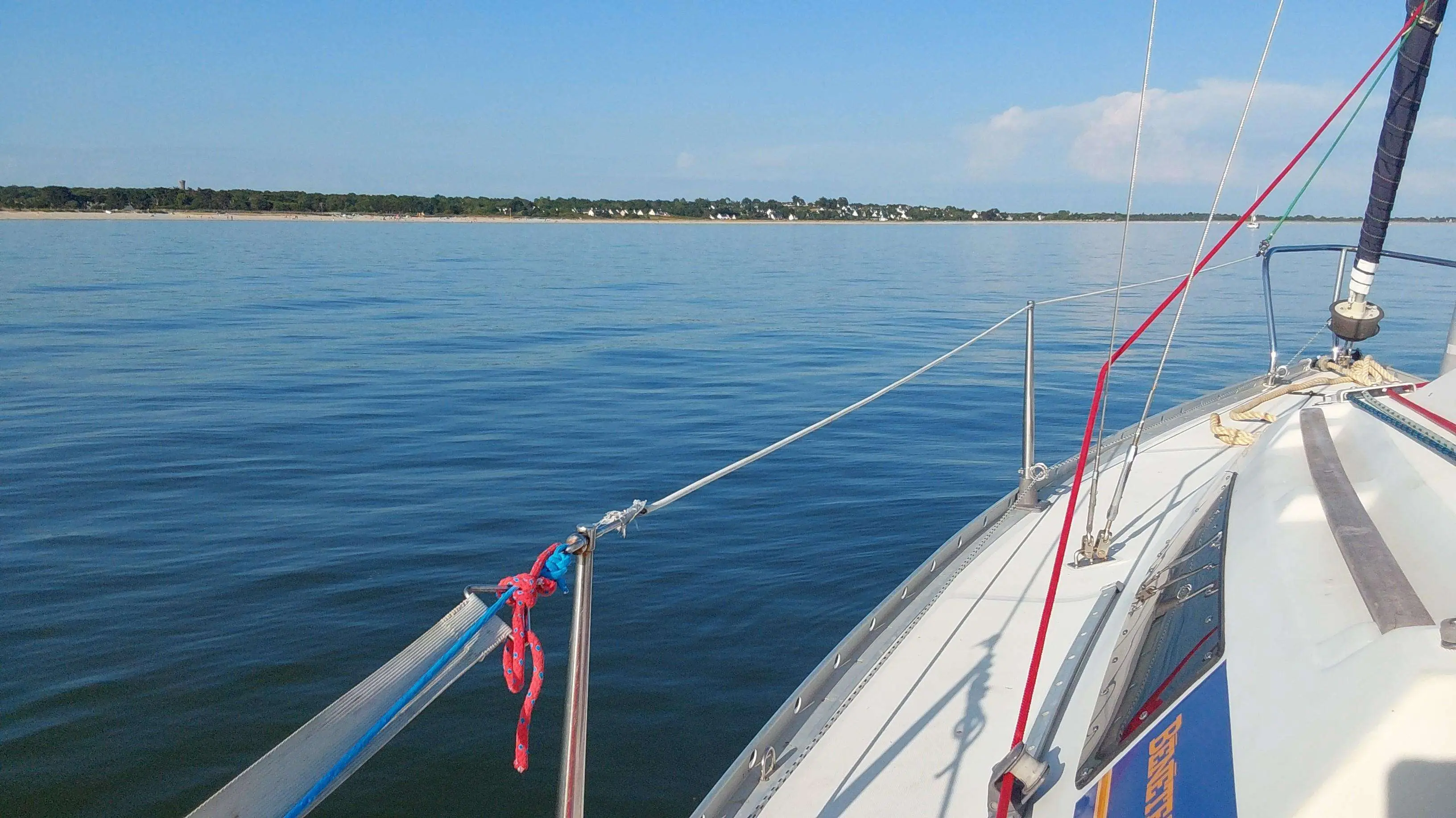 Beneteau First 235 — Vue du pont avant d'un voilier sur une eau calme, avec le rivage en arrière-plan.