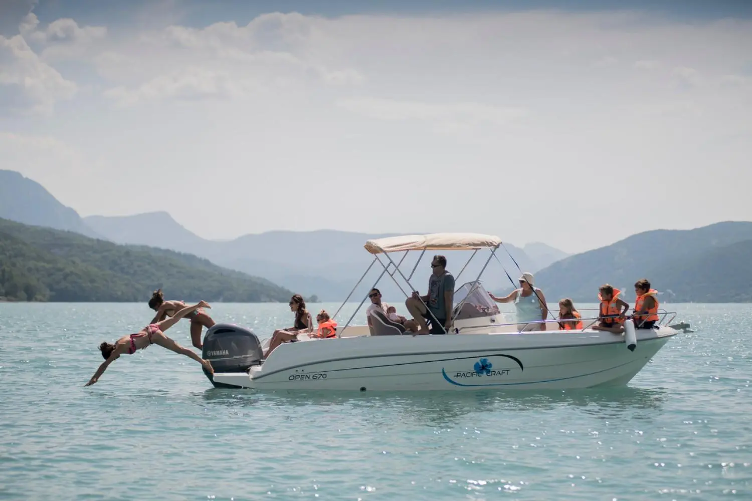 Pacific Craft 670 Plaisance 200cv — Bateau à moteur vu de profil sur un lac, avec plusieurs passagers et des montagnes en arrière-plan.