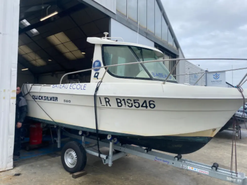Quicksilver 560 — Bateau à cabine blanche sur remorque devant un hangar, vu de trois quarts avant.