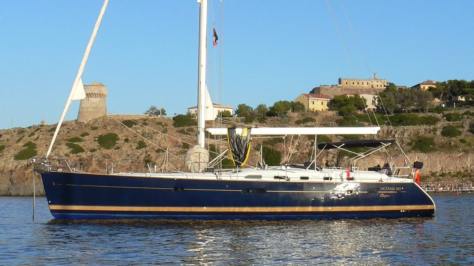 Beneteau Oceanis 523 — Voilier bleu vu de profil sur l’eau, devant une côte rocheuse et des bâtiments historiques.