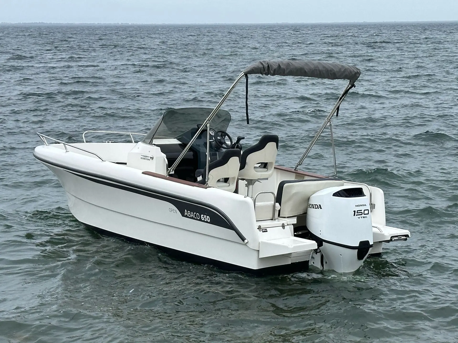 Ocqueteau Abaco 650 Open — Bateau à moteur blanc vu de trois quarts sur l’eau, avec console centrale et taud.