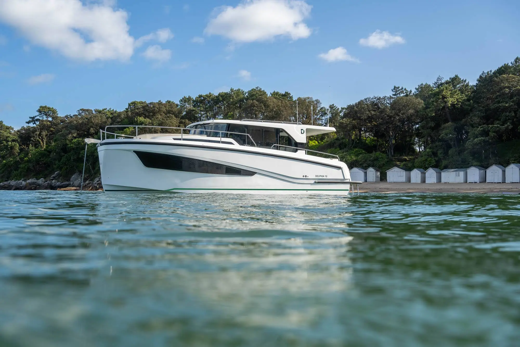 Delphia 10 Sedan — Bateau à moteur blanc vu de profil sur l’eau, devant une plage boisée.