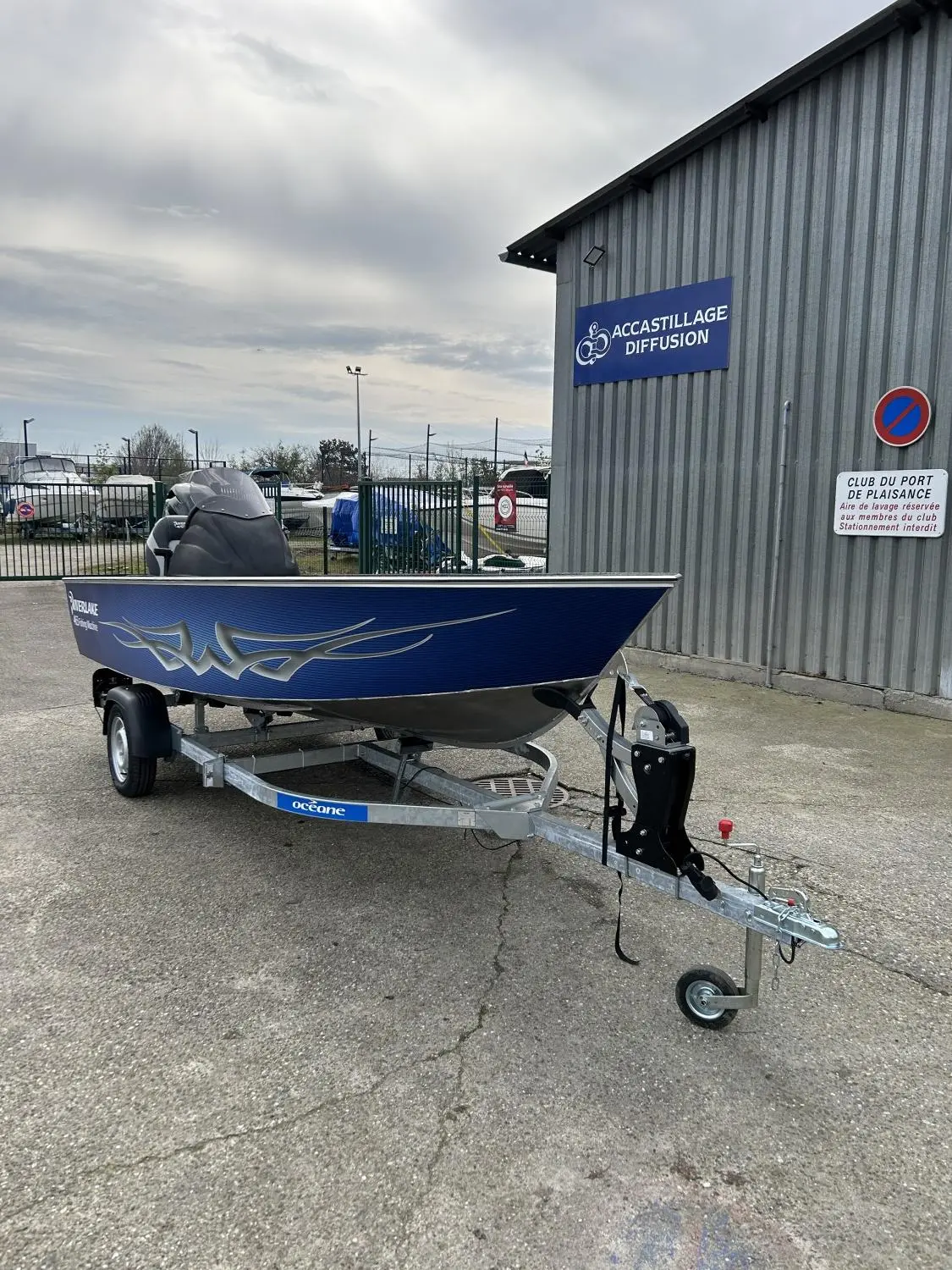 Riverlake 465 Fishing Machine — Petit bateau à moteur bleu et gris sur remorque devant un bâtiment portuaire.