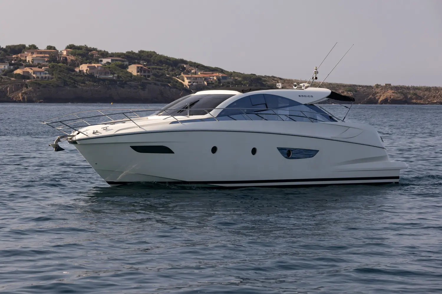 Sogica 47 Openbridge — Bateau à moteur blanc vu de profil sur l’eau, avec une côte habitée en arrière-plan.