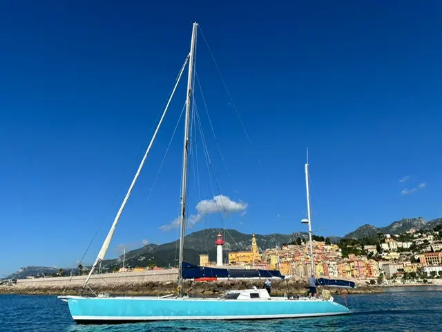 Garcia Eclipse 60 — Voilier turquoise vu de profil devant une digue et une ville côtière.