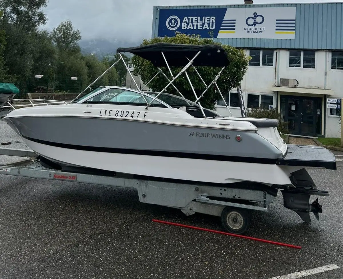Four Winns Sundowner 215 — Bateau de plaisance blanc photographié de profil sur sa remorque, avec taud noir.