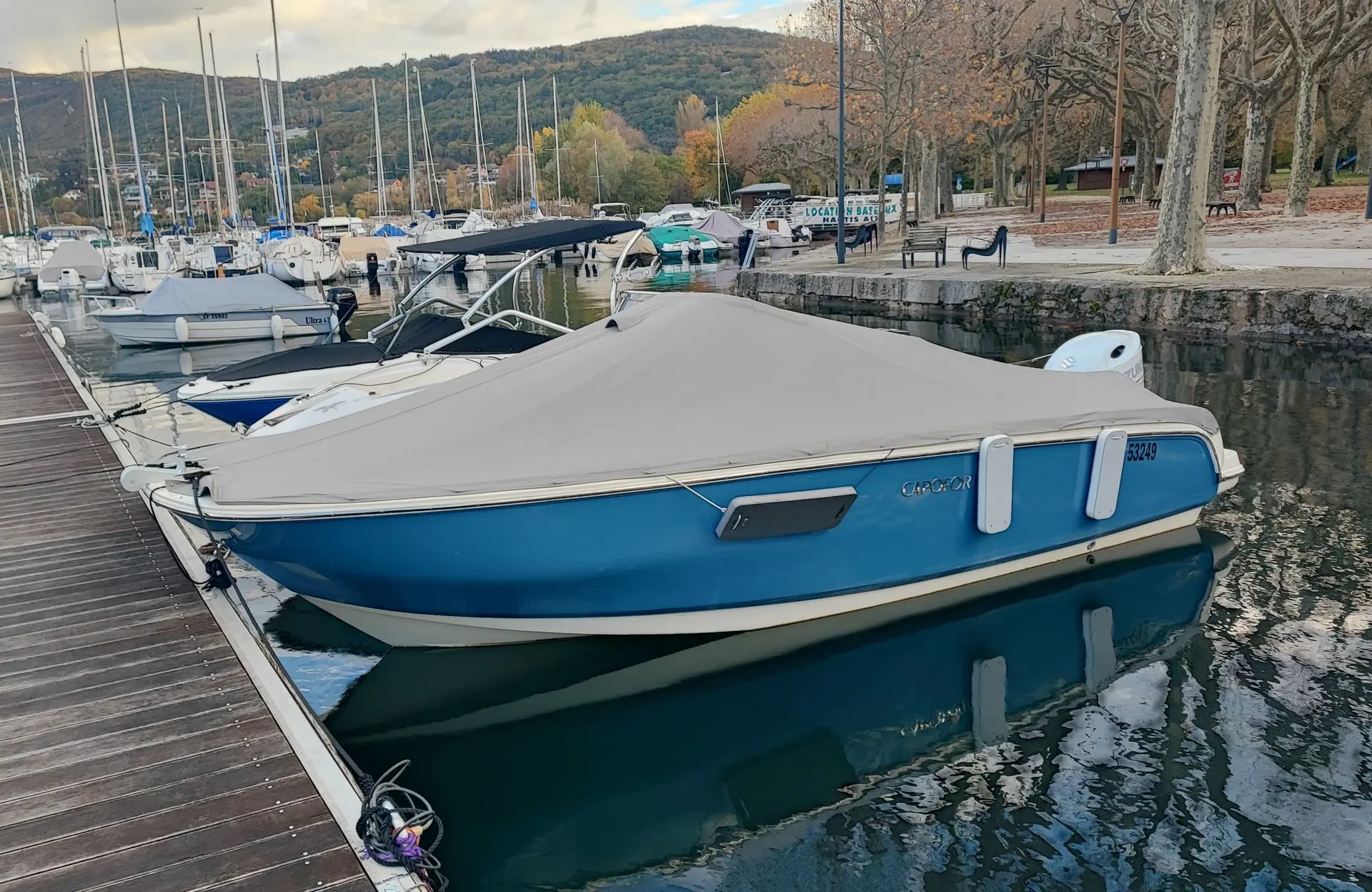 Invictus Capoforte FX 200 — Bateau bleu bâché amarré dans un port, avec des voiliers et des collines en arrière-plan.
