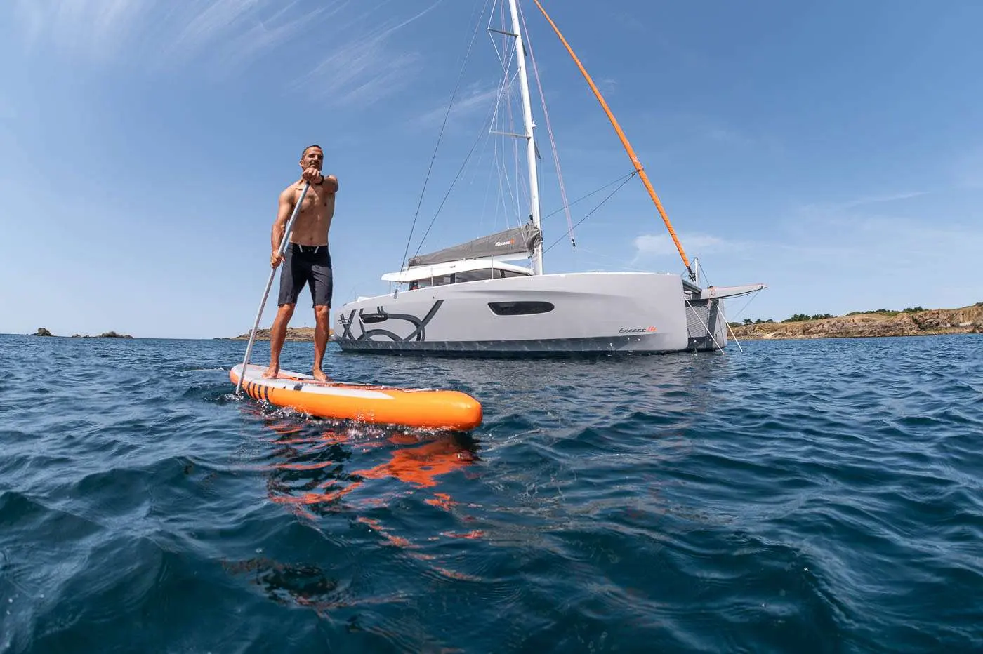 Excess Catamarans Excess 14 — Catamaran à voile vu depuis la mer, avec un homme debout sur une planche.