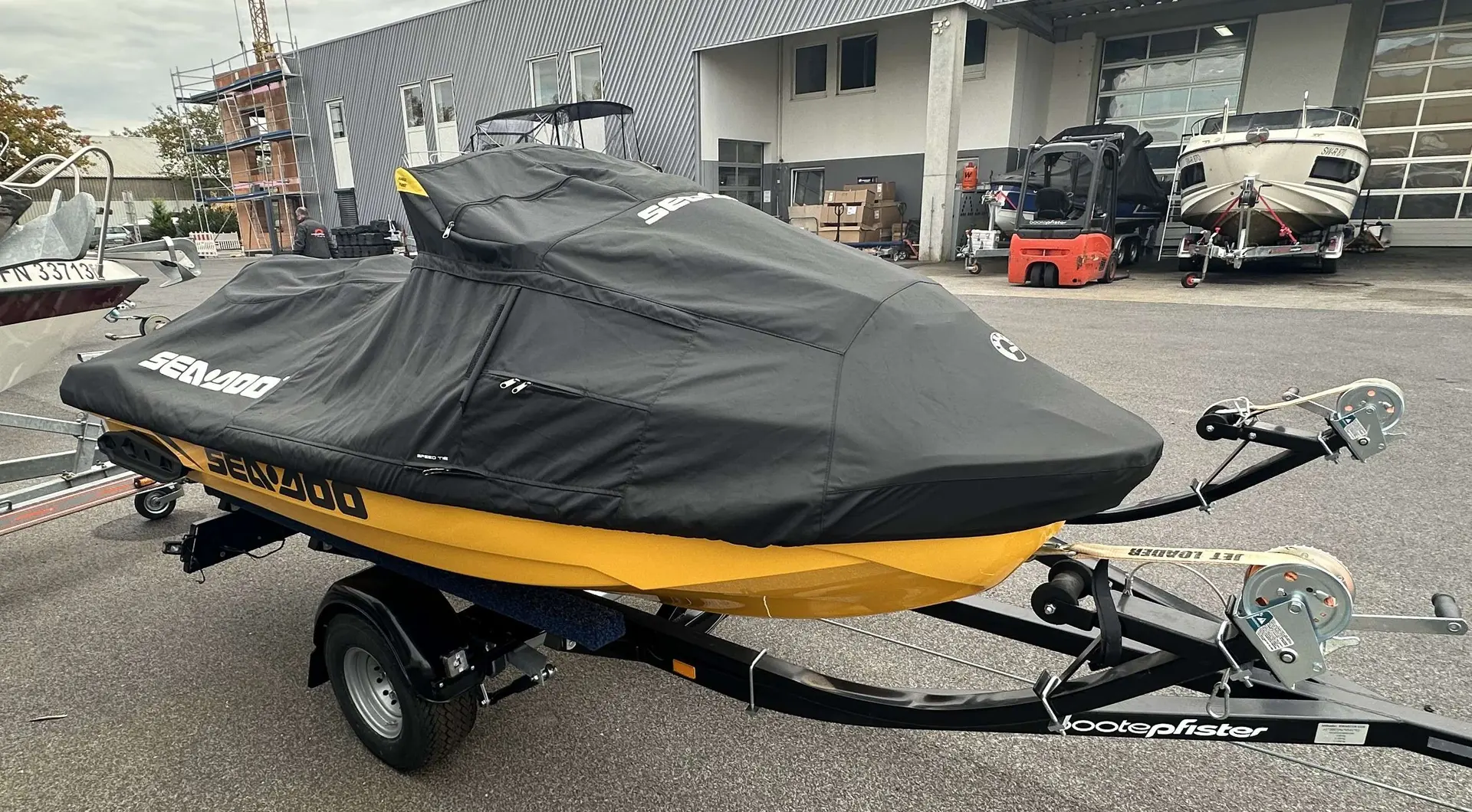 Sea Doo RXT-X 300 — Bateau jaune bâché sur une remorque, devant un bâtiment industriel.