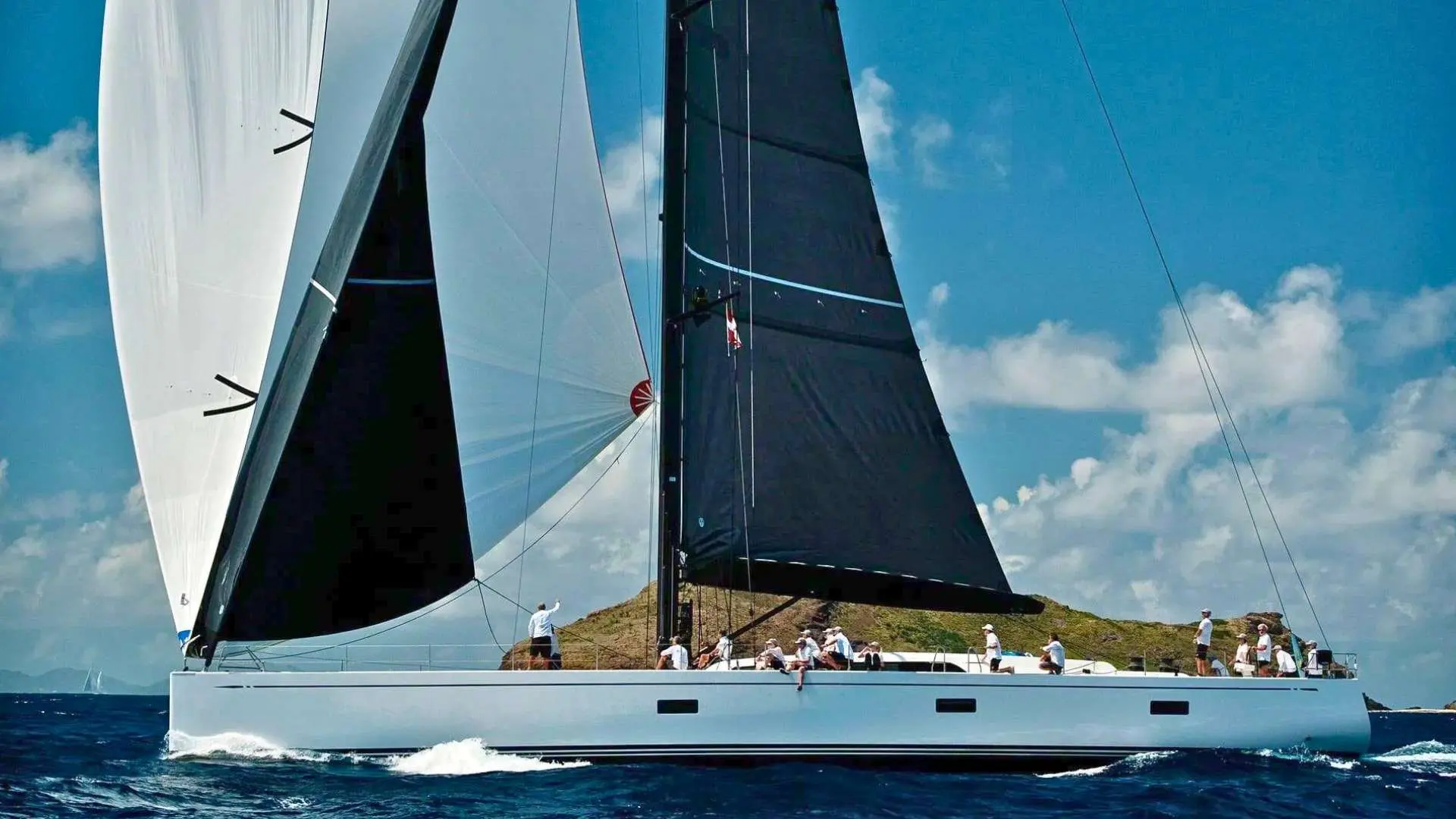 Nautor Swan Swan 98 — Voilier blanc à deux voiles noires naviguant près d’une île sous ciel nuageux.