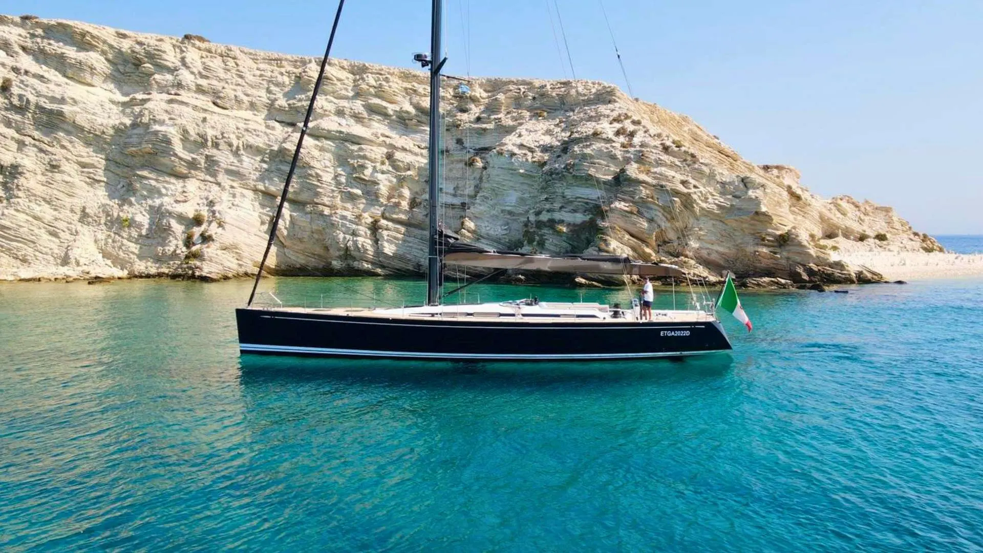Nautor Swan Swan 60 — Voilier noir au mouillage devant des falaises calcaires, dans une eau turquoise.