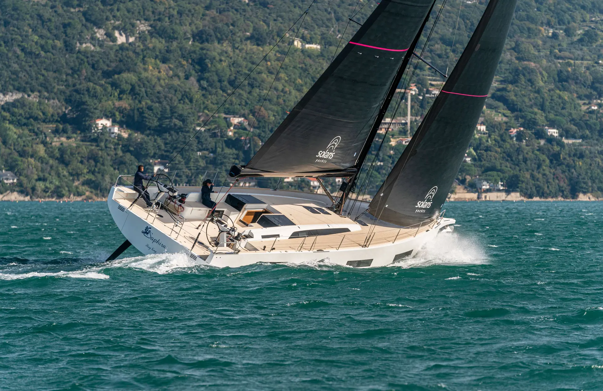 Solaris 55 — Voilier blanc naviguant au près sous voiles noires, avec une côte boisée en arrière-plan.
