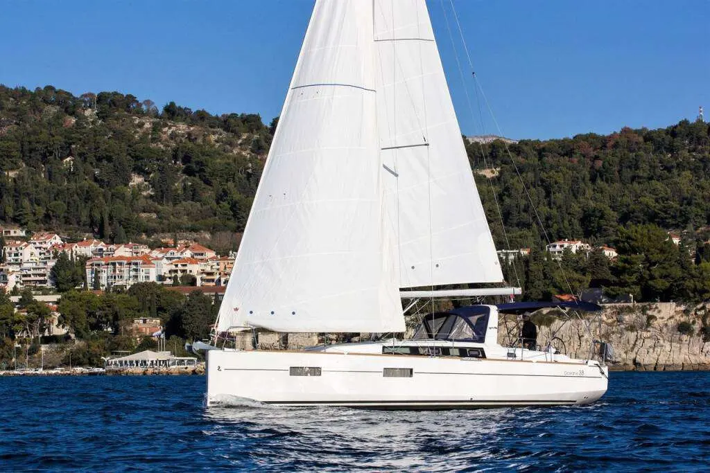 Beneteau Oceanis 38 — Voilier blanc vu de profil, toutes voiles hissées, naviguant près d’une côte boisée.