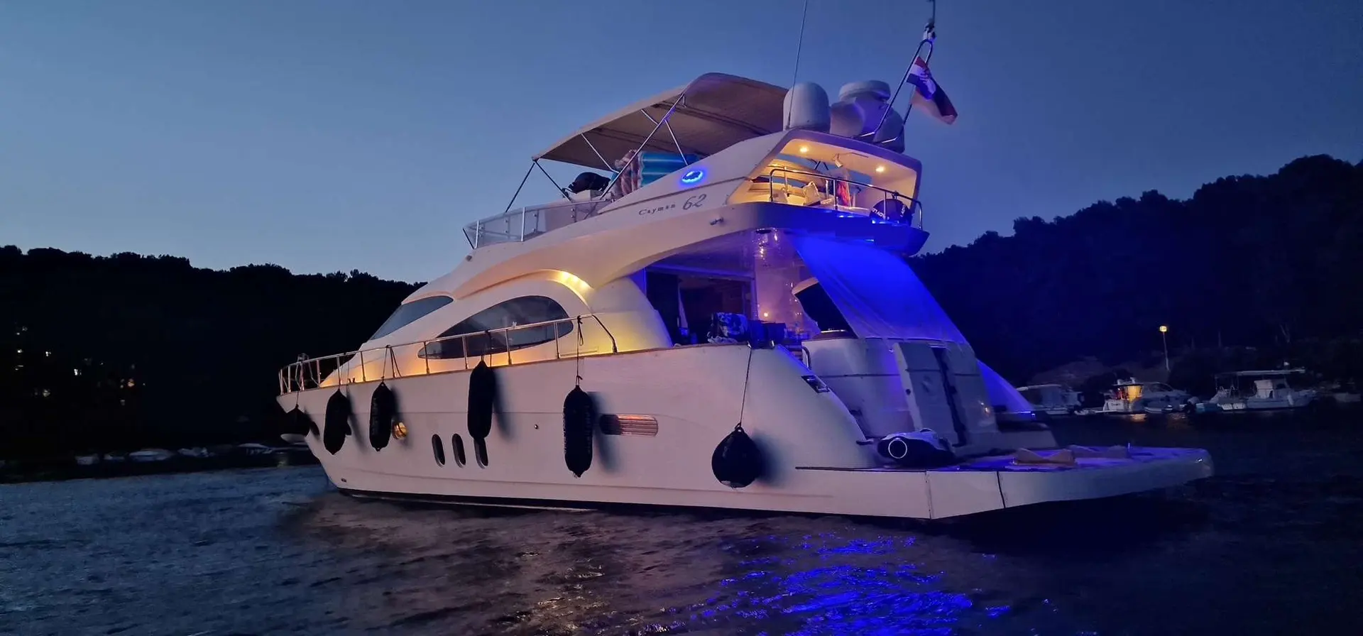 Cayman 62 Fly — Vue arrière trois-quarts d’un yacht blanc éclairé, amarré sur l’eau au crépuscule.