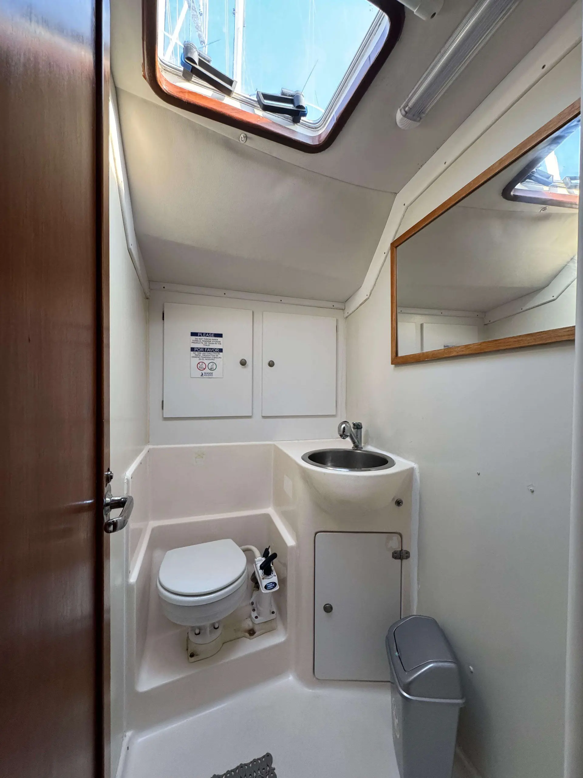 Bavaria 47 — Salle d'eau avec WC, lavabo, miroir et hublot ouvert.