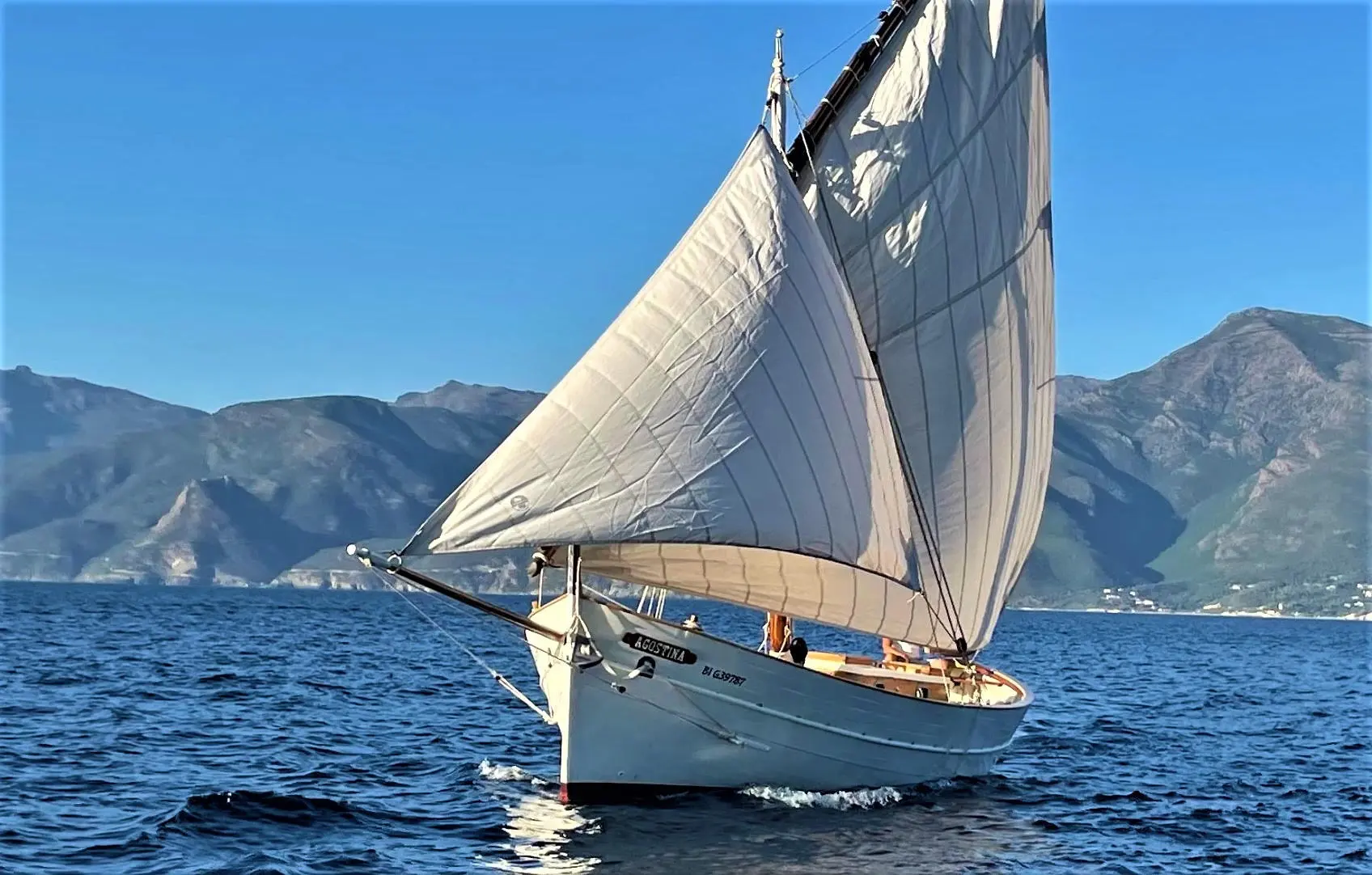 Cantiere Sangermani Rivaneto Ligure — Voilier classique sous voiles naviguant devant un paysage montagneux.
