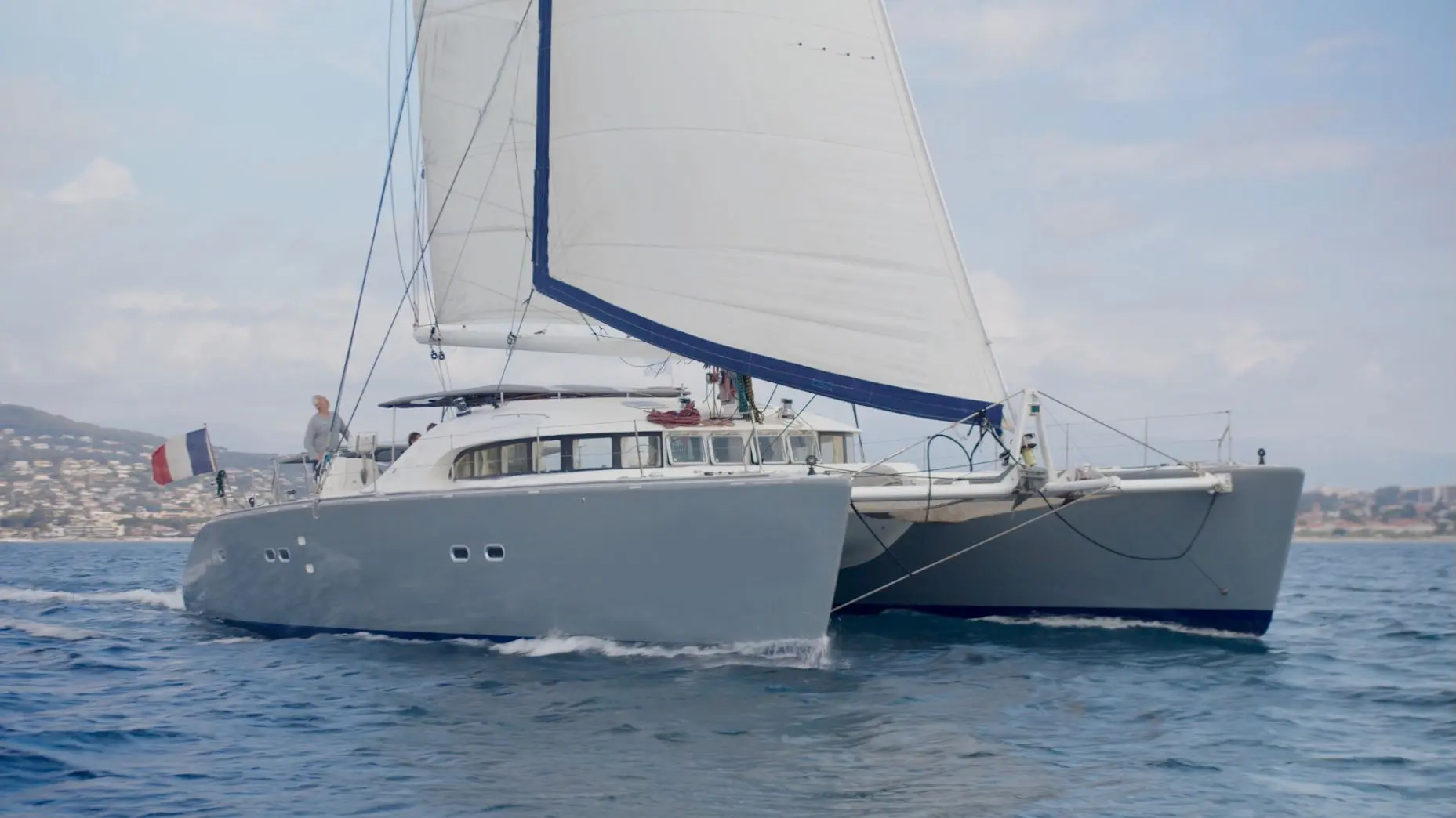 Cnb LAGOON 570 — Catamaran à voile gris naviguant près d'une côte urbanisée.