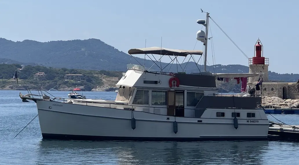 Grand Banks MY 42 — Vue latérale d’un bateau à moteur amarré devant un phare, avec des collines en arrière-plan.