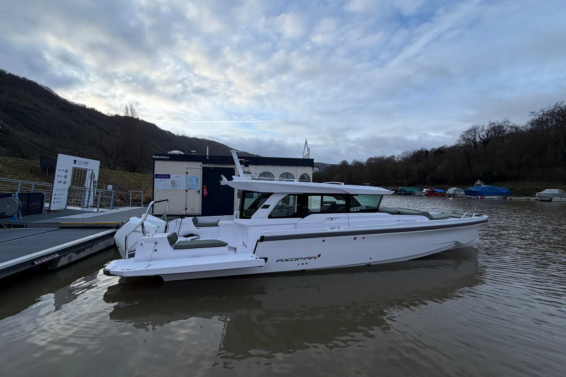 Axopar 38 XC Cross Cabin — Bateau blanc vu de profil, amarré sur un plan d’eau devant une berge boisée.