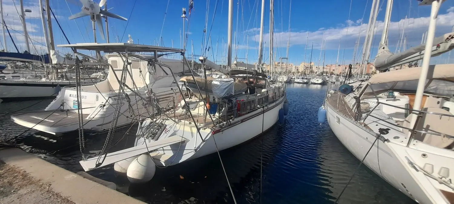 Amel Santorin Ketch — Voilier blanc amarré dans un port, entouré d'autres bateaux à voiles.