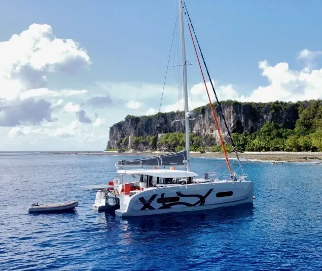Beneteau Excess 11 — Catamaran blanc au mouillage devant une côte rocheuse, avec son annexe à proximité.