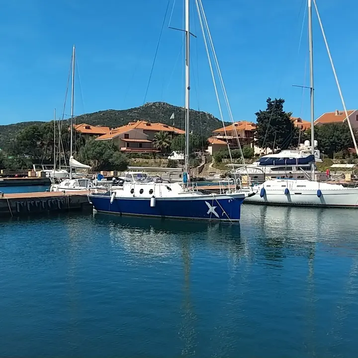 Meta Petit Monde — Voilier bleu amarré dans un port, avec des maisons et une colline en arrière-plan.