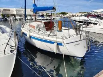 Jeanneau Aquila — Voilier blanc à bande bleue amarré dans un port, vu de trois quarts arrière.
