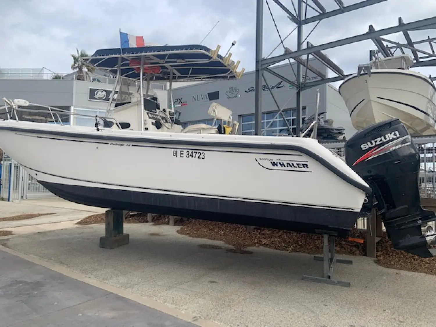 Boston Whaler 26 Outrage — Vue latérale d’un bateau à moteur blanc exposé sur bers, avec moteur hors-bord.