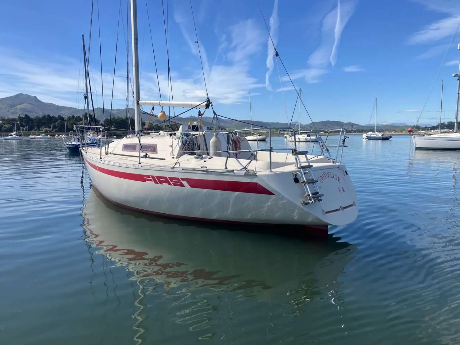Beneteau First 30 — Voilier blanc à bande rouge amarré dans une marina, vu de trois quarts arrière.
