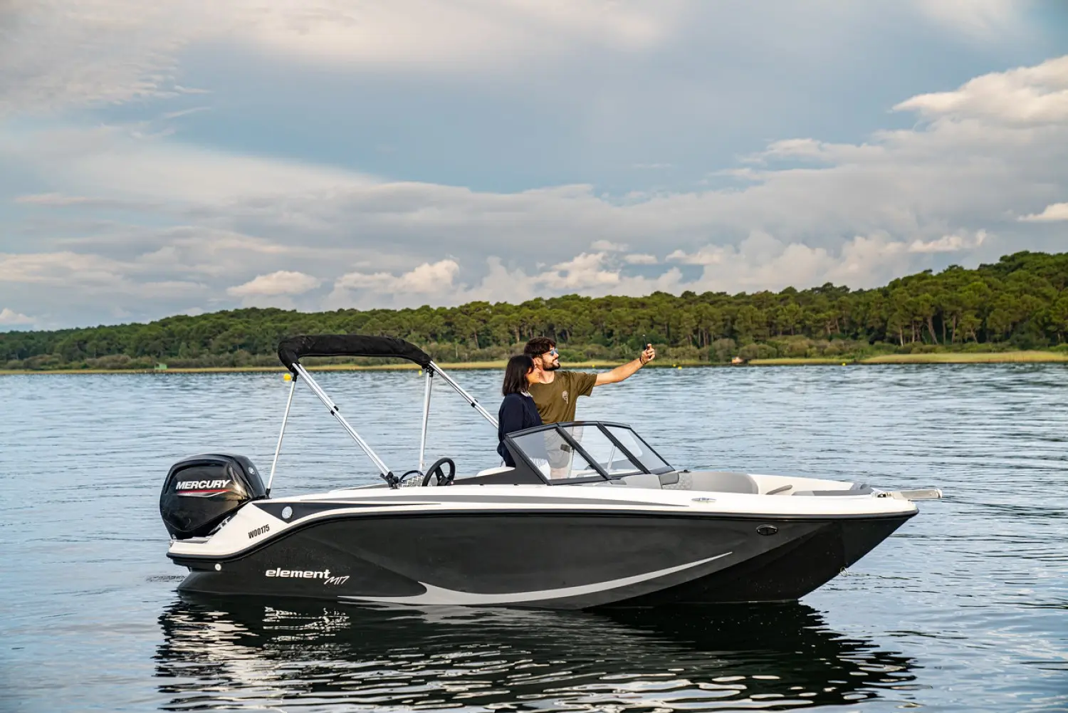 Bayliner Element M17 — Bateau à moteur noir et blanc photographié de profil sur un plan d’eau, avec deux personnes à bord.