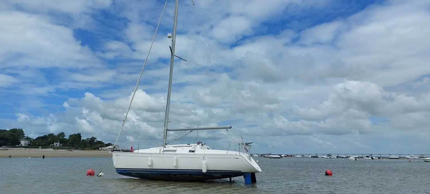 Dufour 30 — Voilier blanc au mouillage dans une baie, avec le rivage et des bateaux en arrière-plan.