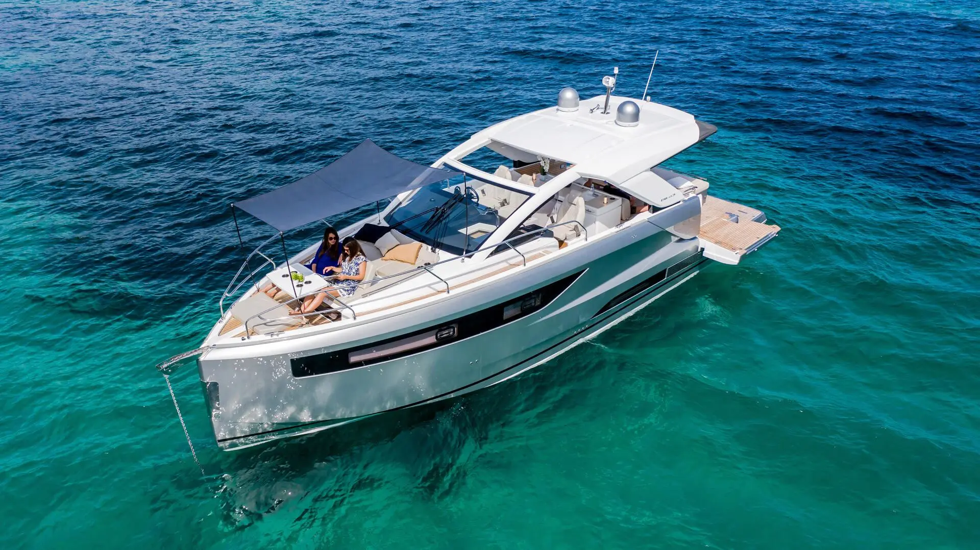 Jeanneau DB 43 — Bateau à moteur blanc vu de trois-quarts sur une mer turquoise, avec deux personnes à bord.