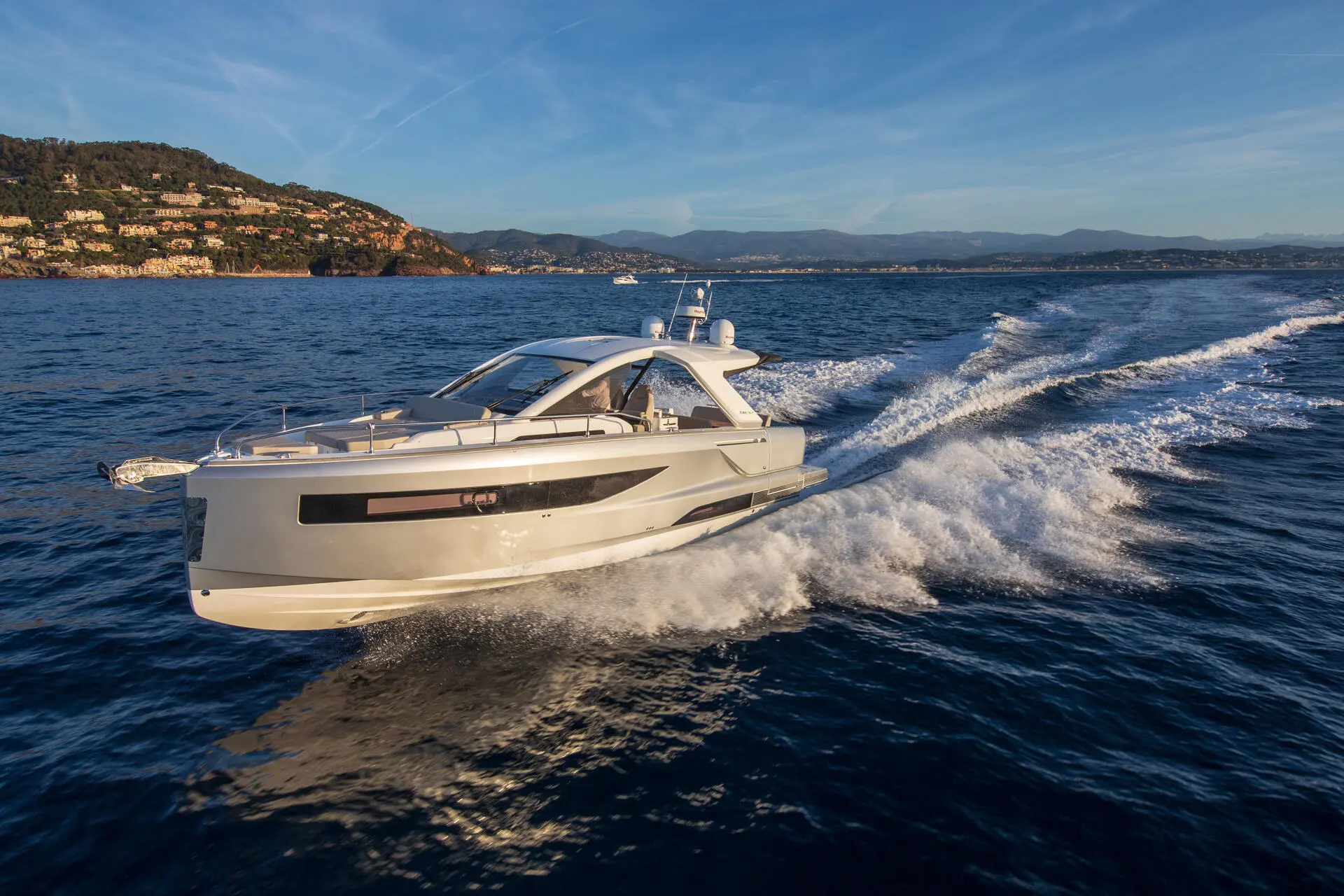 Jeanneau DB 37 — Bateau à moteur vu de trois quarts naviguant au large d’une côte montagneuse.