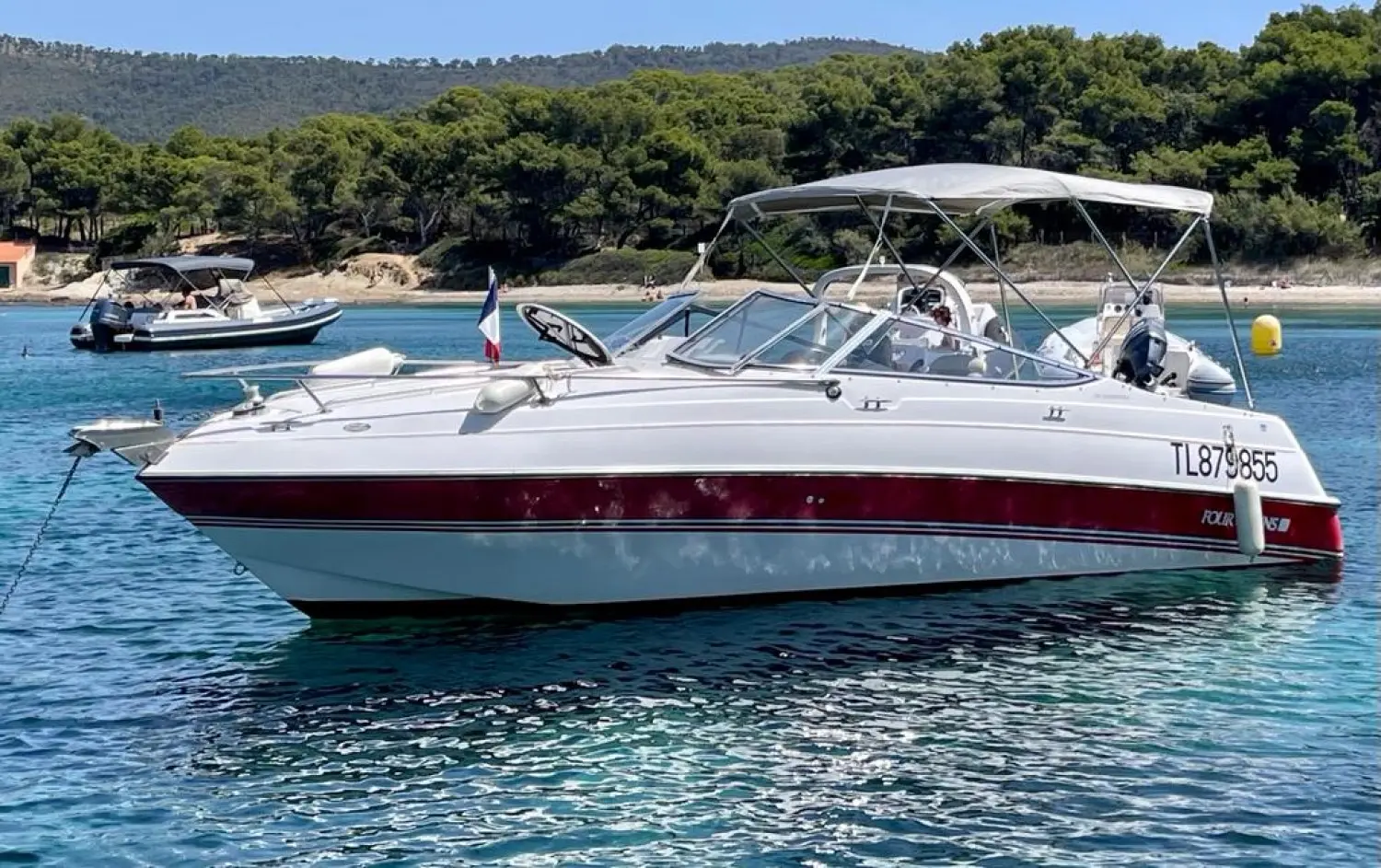 Four Winns Sundowner 235 — Bateau à moteur blanc et rouge amarré sur une eau calme, vu de trois quarts.