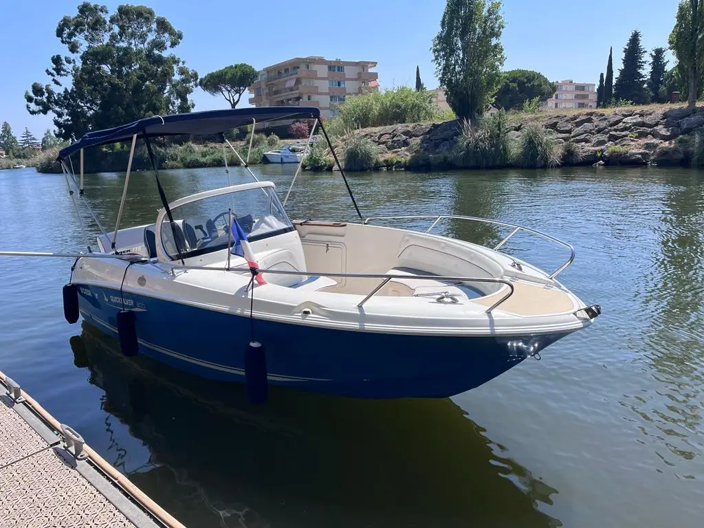 Quicksilver 600 Commander — Bateau à moteur blanc et bleu amarré au bord d’une voie d’eau.